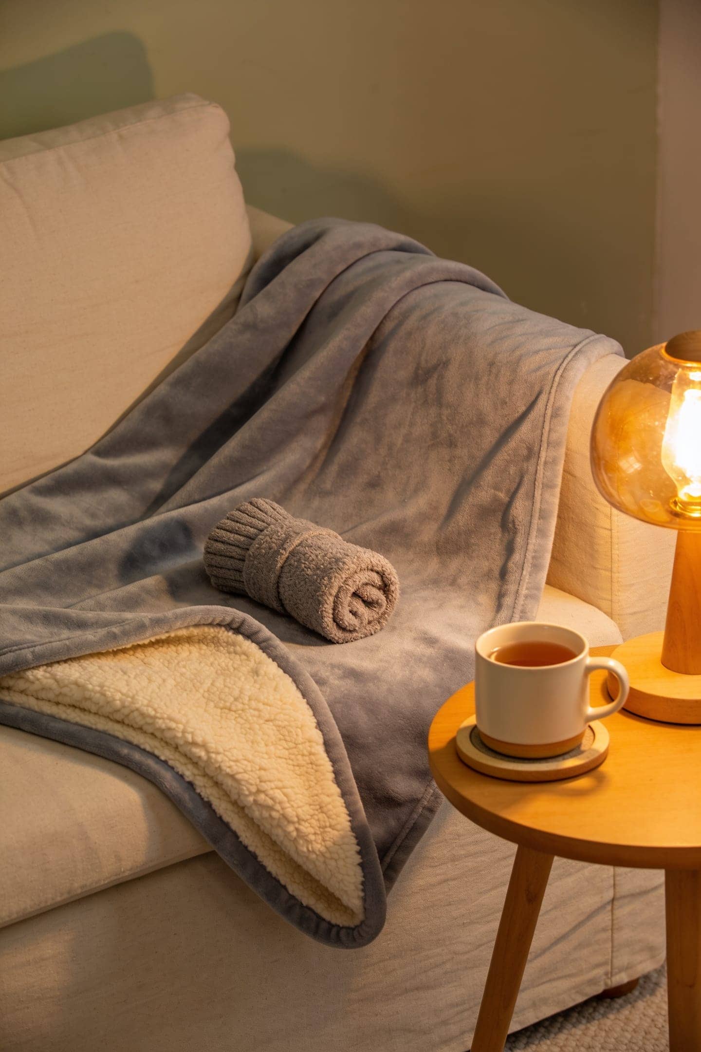 a grey heated throw, a reading pillow and a mug on a warming plate on a sofa in soft evening lamplight